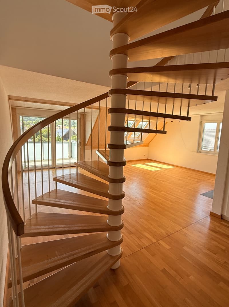 Spiral staircase with wooden steps and a white central column, leading to an open area with wooden flooring and large windows.