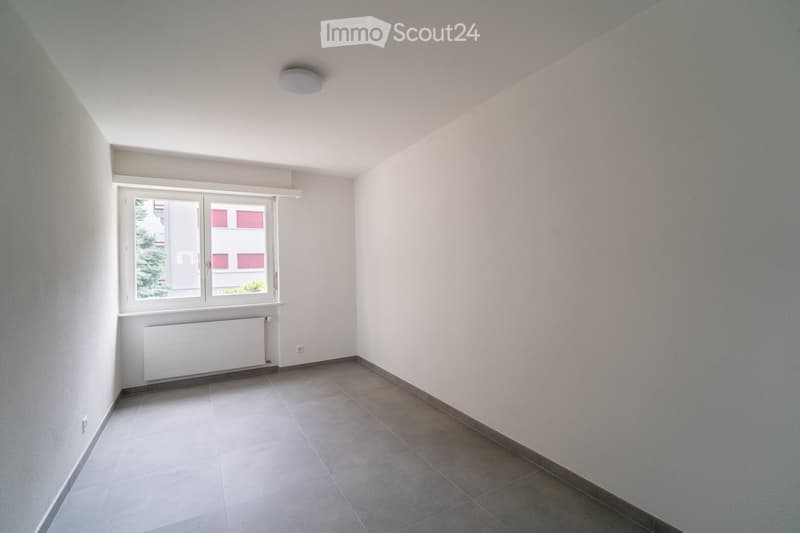 Empty room with tiled floor, white walls, a window with white frame, radiator, and a ceiling light fixture.