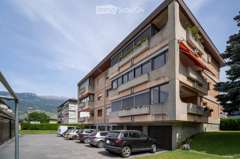 Modern multi-story residential building with balconies, large windows, and a parking area with several cars.