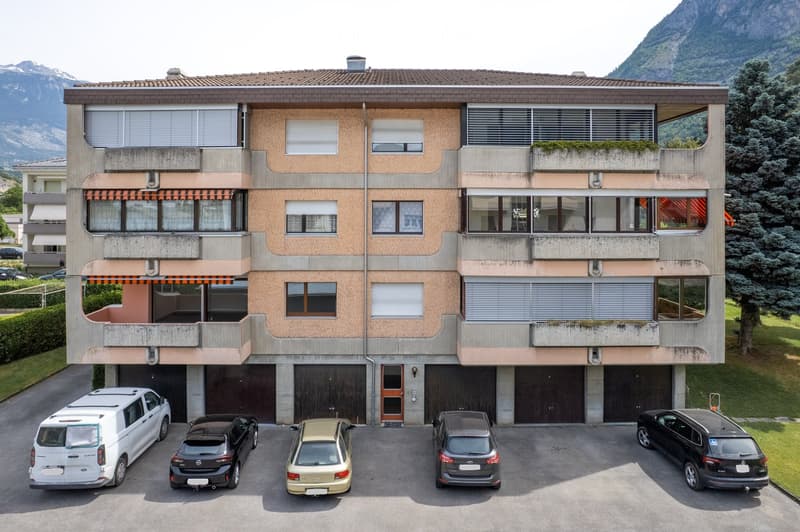 Modern multi-story building with balconies, large windows, and a garage area with several parked cars.