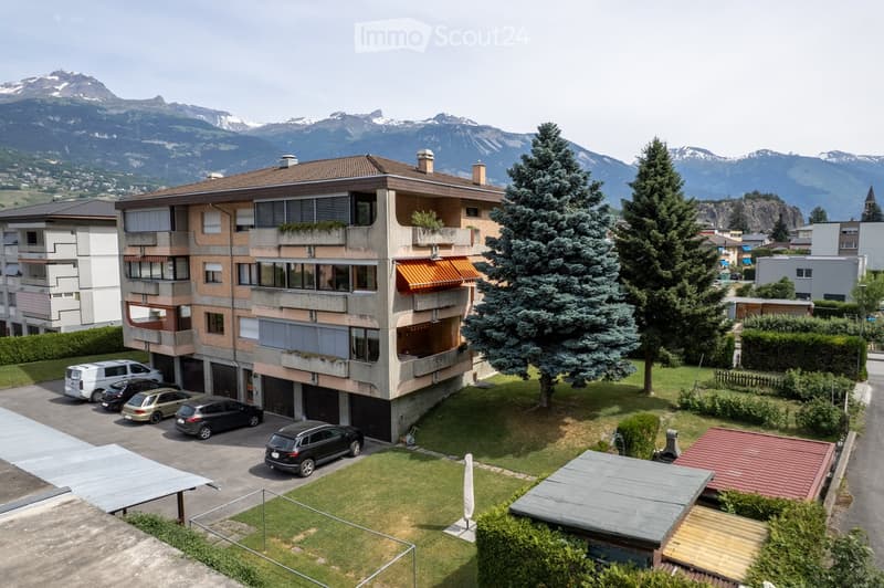 Multi-story residential building with balconies, surrounded by greenery and parked cars, with mountain views in the background.