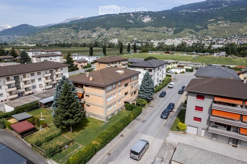 Aerial view of a residential area with multiple apartment buildings, surrounded by greenery and a road with parked cars. Distant mountains and vineyards are visible in the background.