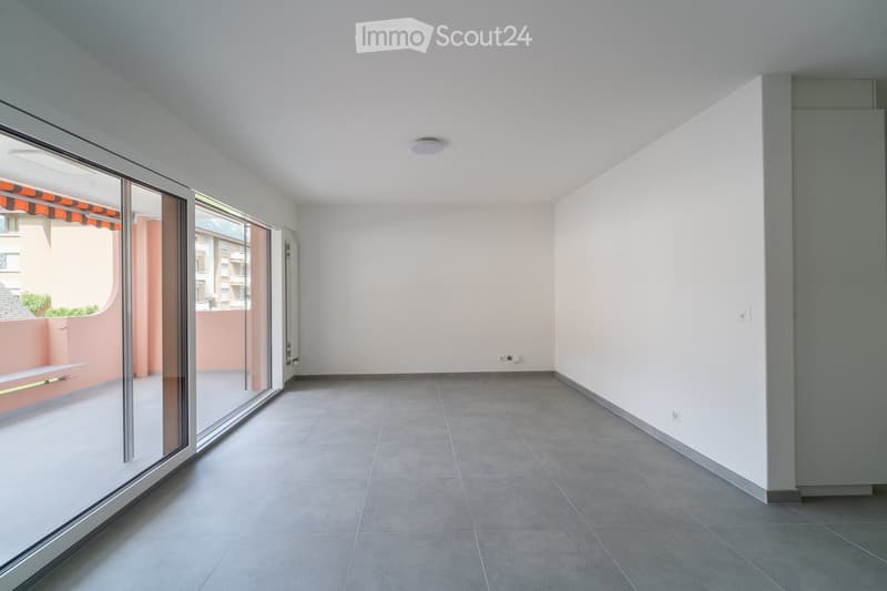Empty room with grey tiled floor, white walls, large glass sliding doors leading to a balcony, and a visible ceiling light fixture.