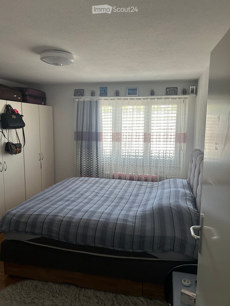 Cozy bedroom with a neatly made bed, a wardrobe, and a window with curtains. A handbag is hanging on the wardrobe, and there are decorative items on the windowsill.