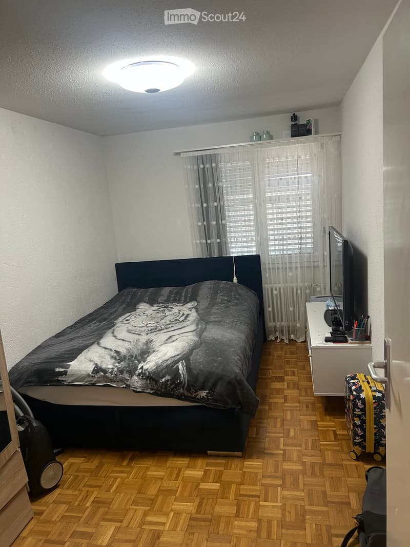 A tidy bedroom with a wooden floor, a bed with a tiger print duvet, a nightstand with a screen, a suitcase, and a radiator.