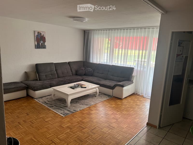 Living room with a grey sectional sofa, white coffee table, and a picture frame on the wall. The room features wooden flooring and large windows with sheer curtains.