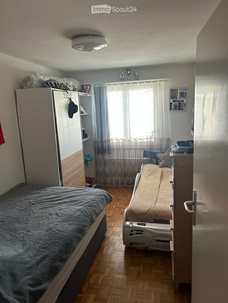 A small bedroom with a double bed, a wardrobe, a chair, a small table, and a window with curtains. The room has wooden flooring and a ceiling light.