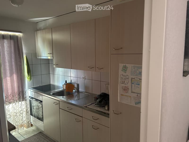 A compact kitchen with light-colored cabinets, a sink, an oven, and a refrigerator. The space includes a towel hanging on the wall and a few magnets on the fridge.