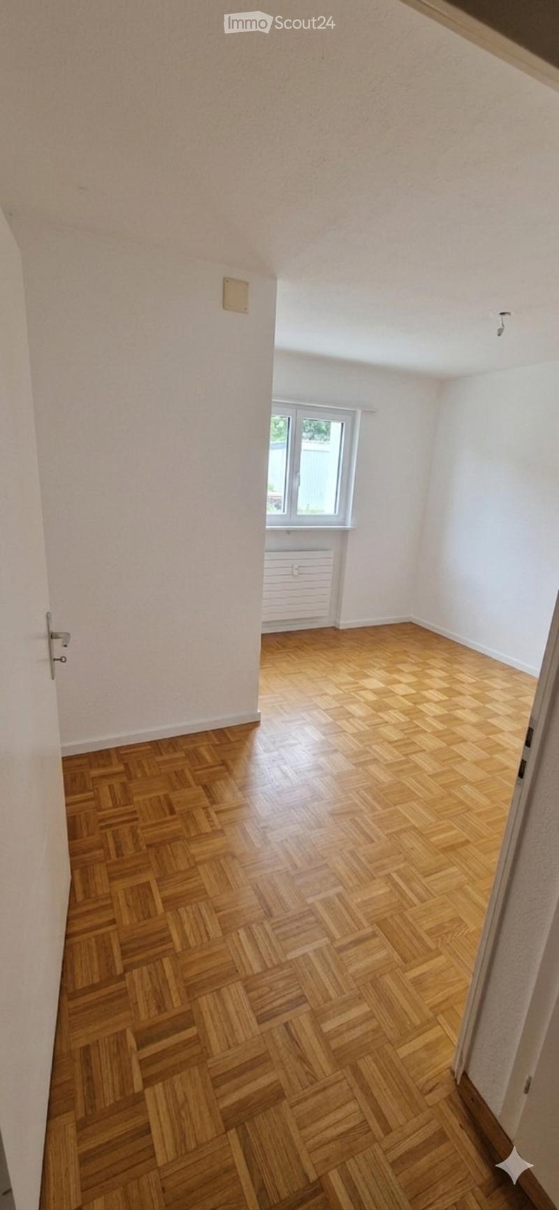 Empty room with wooden herringbone flooring, white walls, and a window with a radiator below it.