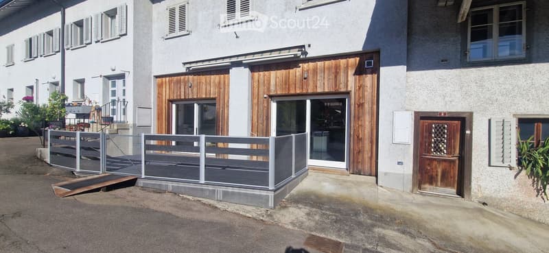 Two-story building with a raised ground floor, featuring a wooden entrance, a fenced balcony area, and multiple windows with shutters.
