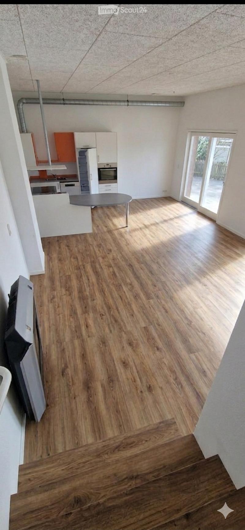 Open-plan living and dining area with wooden flooring, a round dining table, and a small kitchen area with white and orange cabinets, an oven, and a refrigerator.