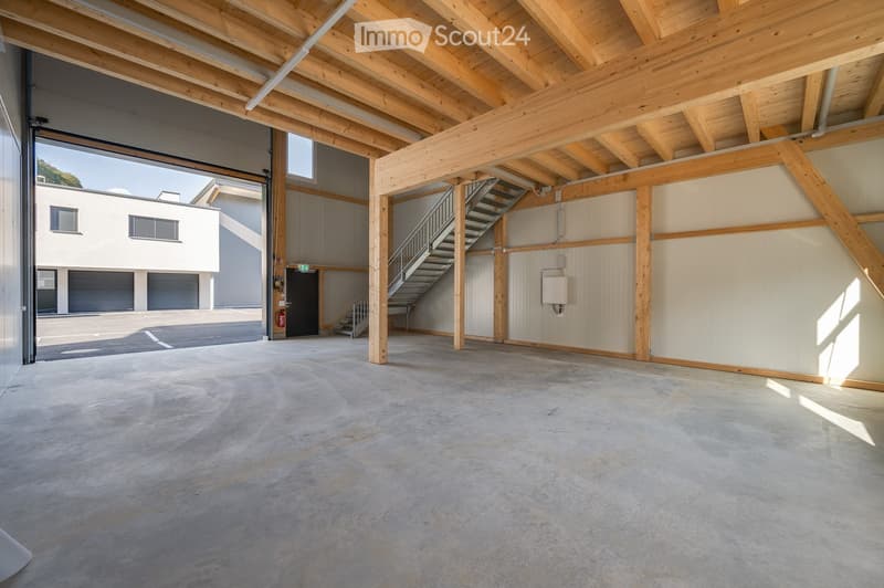 Empty warehouse space with exposed wooden beams, concrete floor, and a staircase leading to an upper level. The space is well-lit with natural light from large windows and has a modern, industrial design.