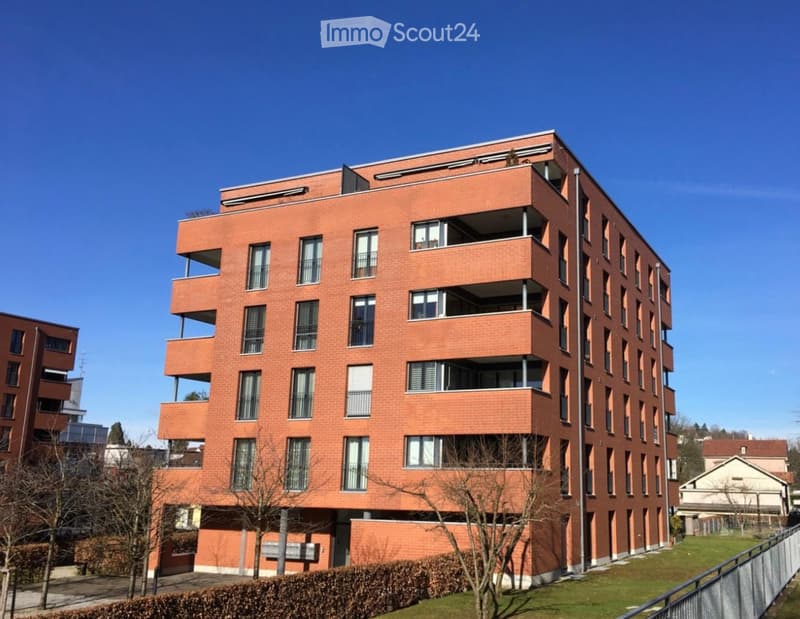 Modern multi-story apartment building with red brick facade, multiple balconies, and large windows.