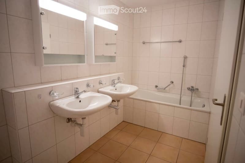 Modern bathroom with two sinks, a bathtub, and two mirrors with built-in storage. The walls and floor are tiled in neutral tones.
