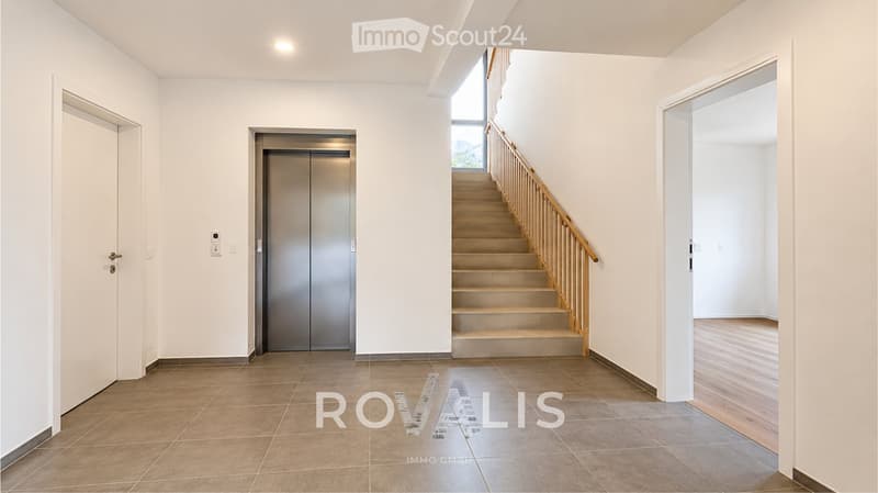 Bright hallway with tiled floor, elevator, staircase with wooden railing, and doors leading to other rooms.