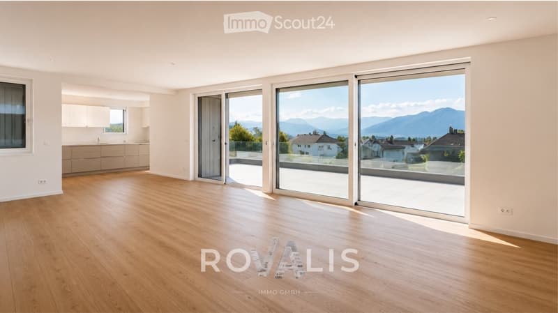 Bright, empty room with wooden flooring, large sliding glass doors leading to a balcony, and a view of mountains and neighboring houses.