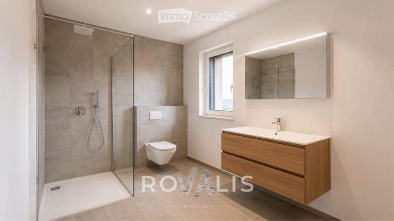 Modern bathroom with a glass-enclosed shower, toilet, wooden vanity with sink, and a large mirror with built-in lighting. The space features neutral tones and a large window providing natural light.