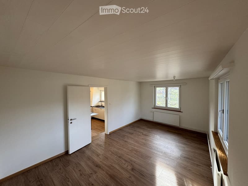 Empty room with wooden flooring, white walls, and large windows. A door leads to another room with a kitchen visible in the background.