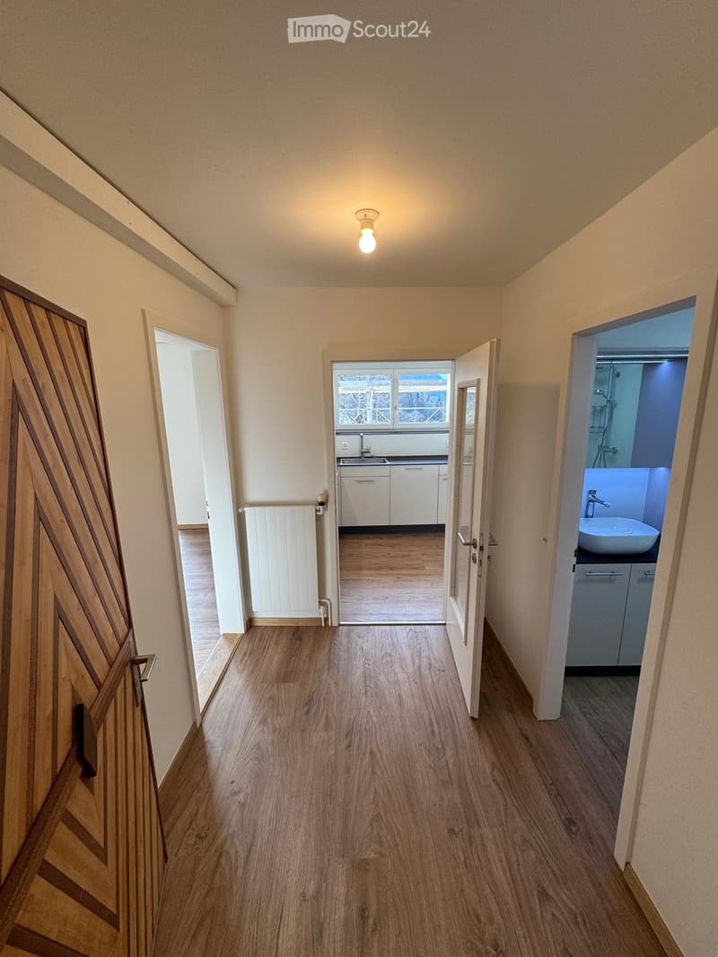 Empty hallway with wooden flooring, white walls, and open doors leading to a kitchen and bathroom. The kitchen has white cabinets and a sink, while the bathroom features a sink and shower area.