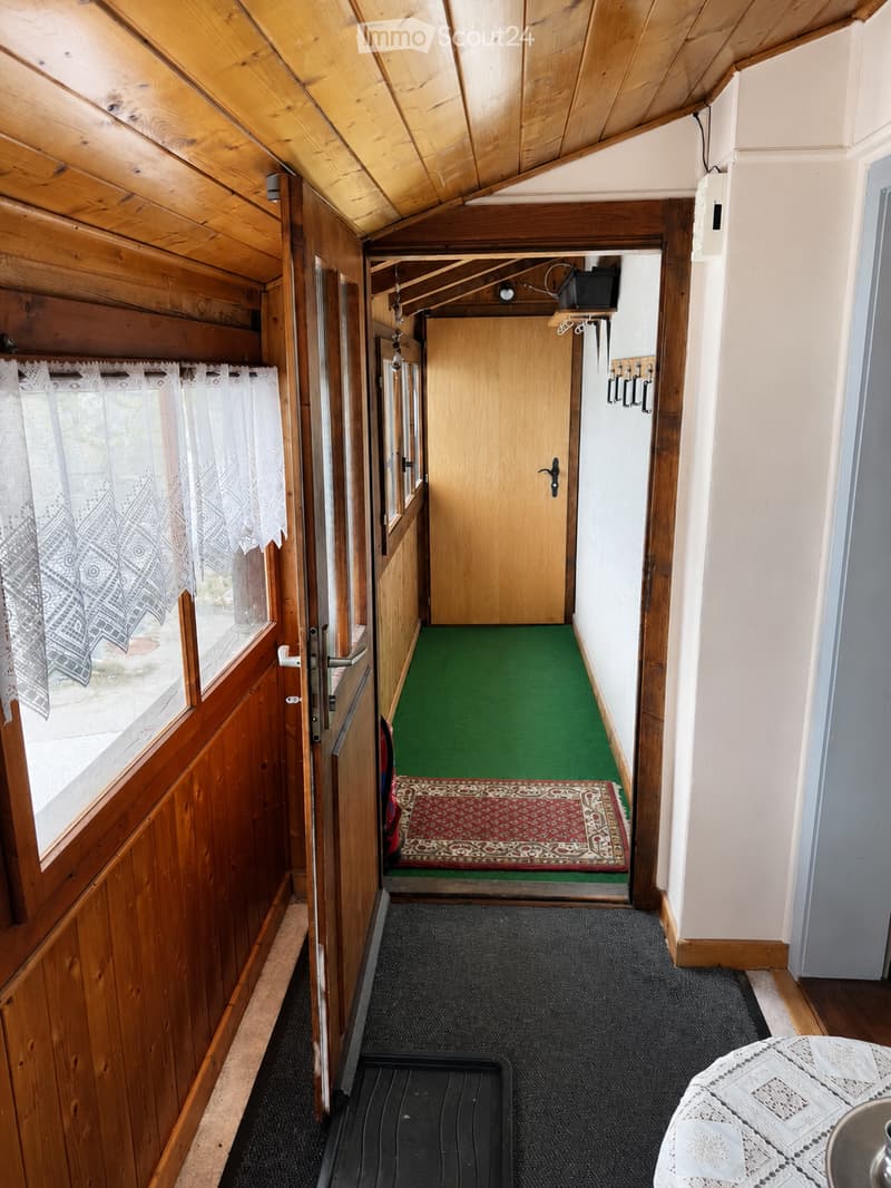 A narrow wooden hallway with green carpeting, a small red and white patterned rug, and a window with white lace curtains on the left side.