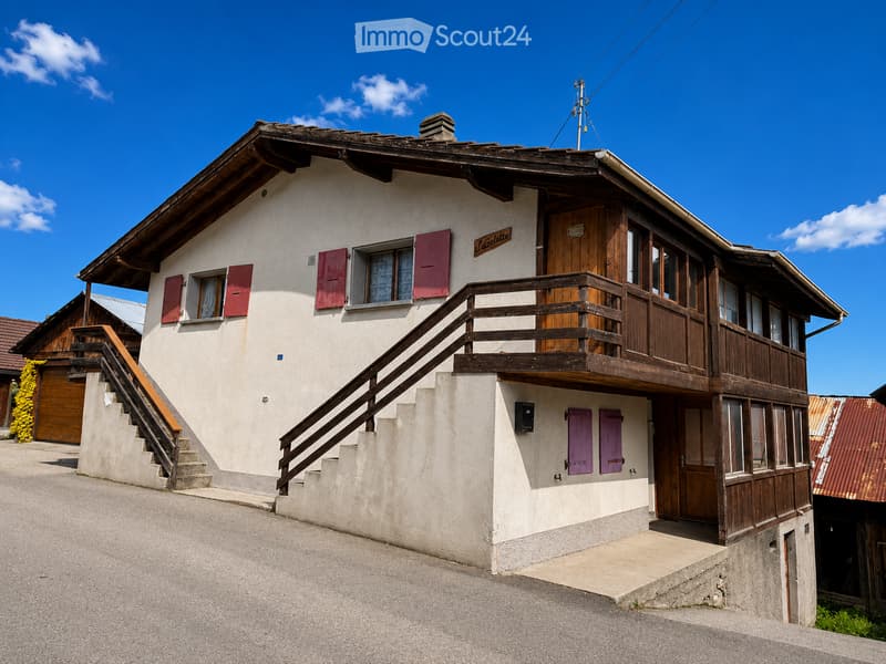 Two-story house with white walls, wooden balcony, red shutters, and a wooden front door. Stairs lead up to the entrance.