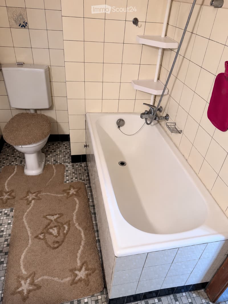 Bathroom with a bathtub, toilet, towel, and bathmat. The walls are tiled, and the floor has a mosaic tile pattern.