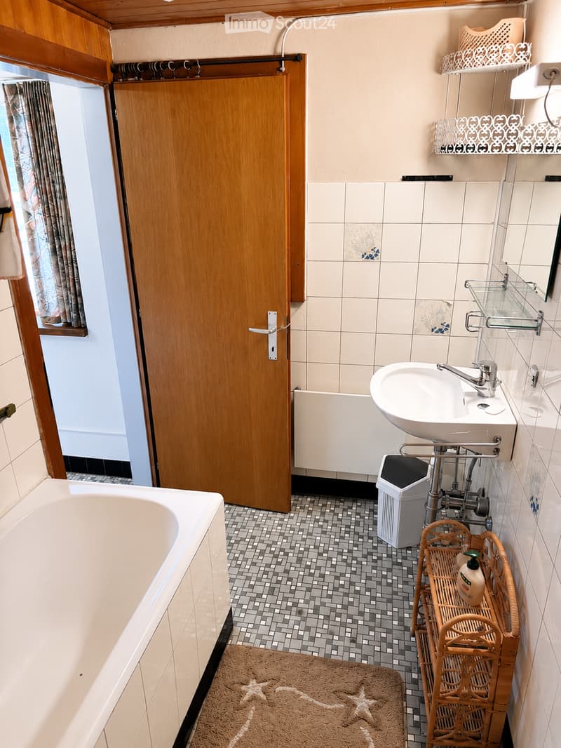 Bathroom with a bathtub, sink, mirror, wicker basket, and a curtain on the window. The floor is tiled with a small rug in front of the bathtub.