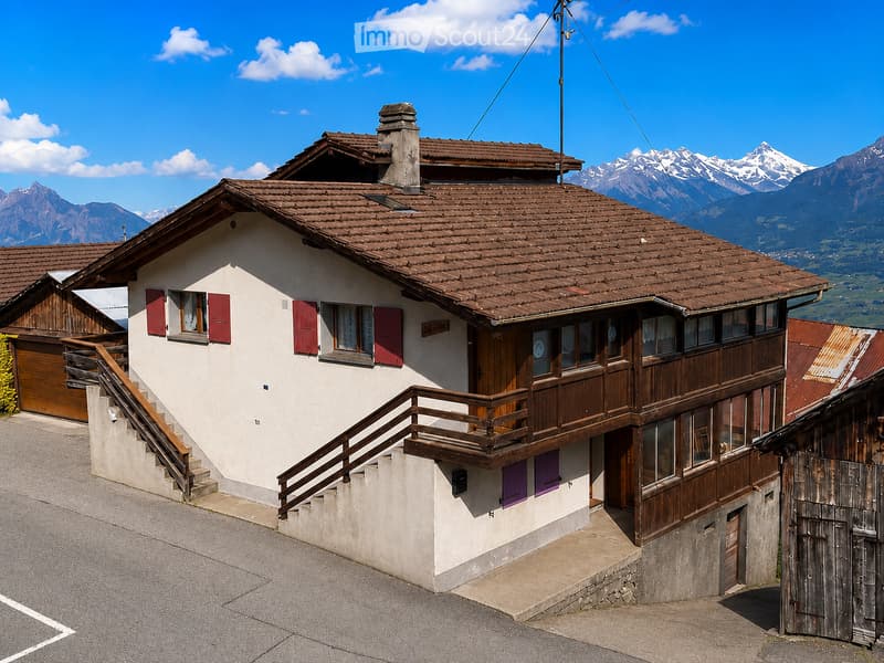 Two-story house with a tiled roof, white walls, red shutters, and a wooden balcony. The house is situated in a mountainous area with a clear blue sky and distant snow-capped peaks.