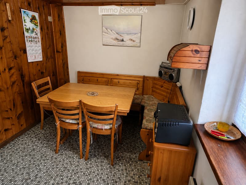 Cozy wooden-paneled room with a round wooden table surrounded by four chairs, a built-in wooden bench with cushioned seating, a small boombox, a printer, and a small shelf with a cup and utensils.