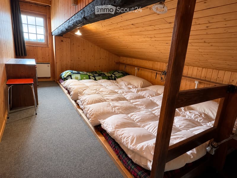 Cozy attic bedroom with wooden walls and slanted ceiling, featuring a large wooden bed with multiple sleeping bags and pillows, a small wooden stool, and a window with curtains.