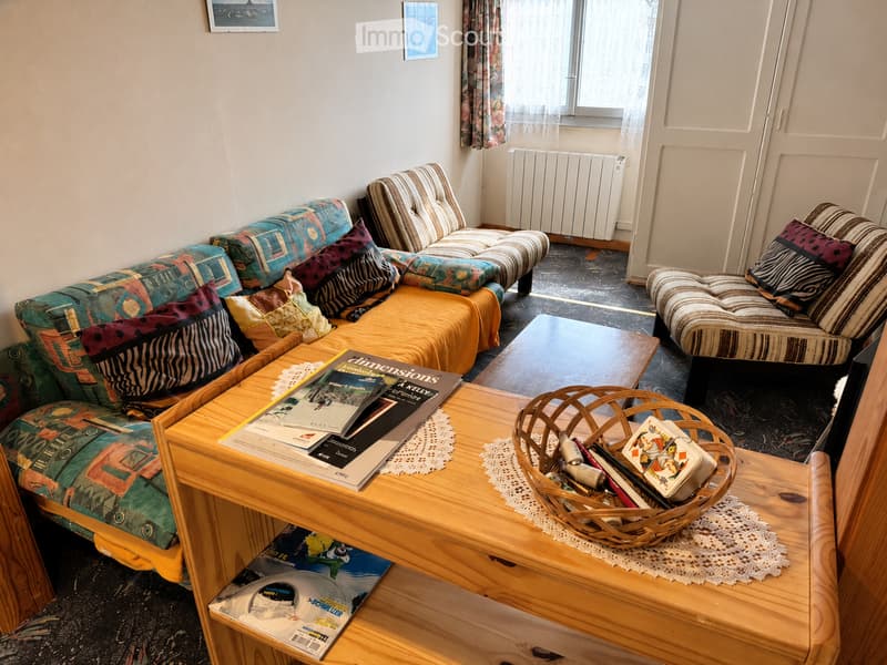 A cozy living room with a colorful sofa, ottoman, coffee table, and a wicker basket on the floor. The room features a radiator, large windows with curtains, and a patterned rug.
