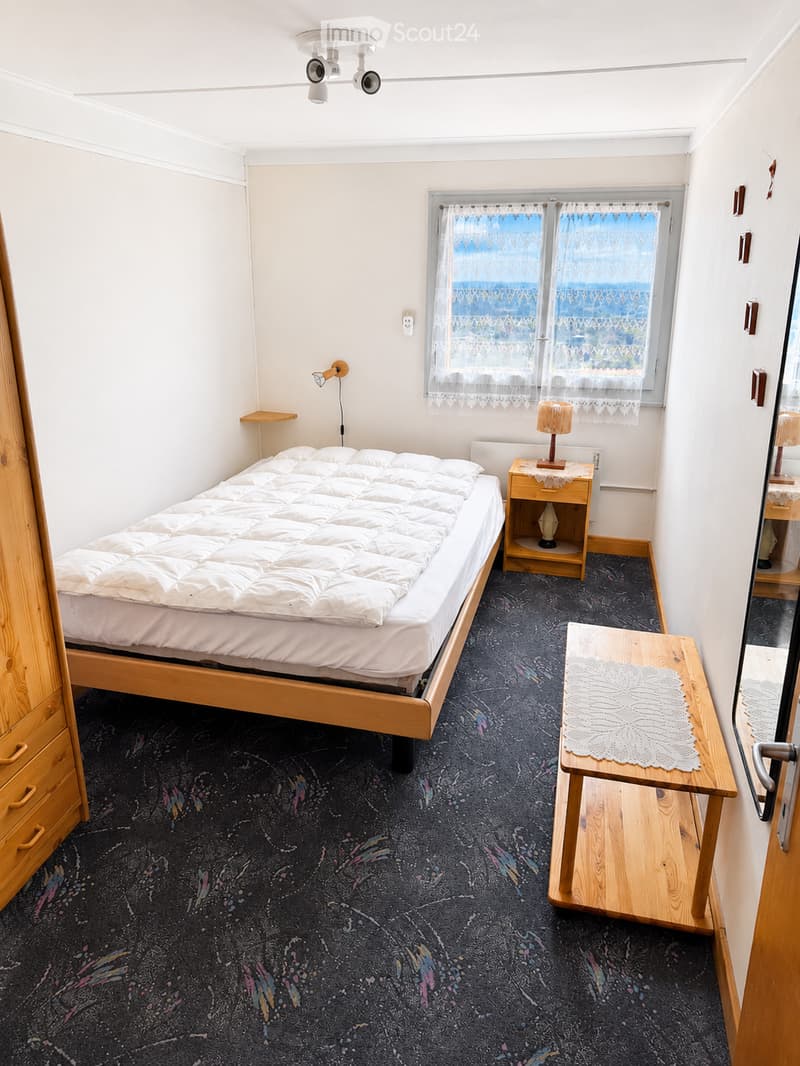 Cozy bedroom with a single bed, nightstand, lamp, bench, and wardrobe. The room features a patterned carpet and large windows with lace curtains.