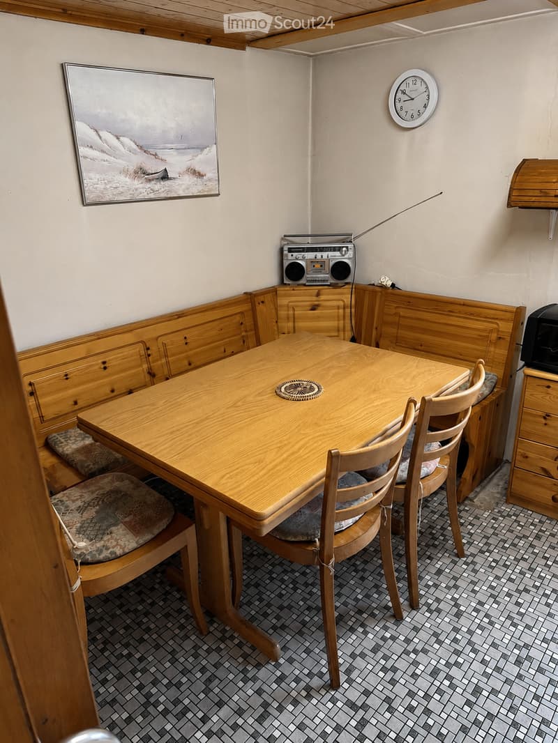 A small, cozy dining area with a wooden table, four chairs, a boombox, a clock, and a picture frame on the wall.