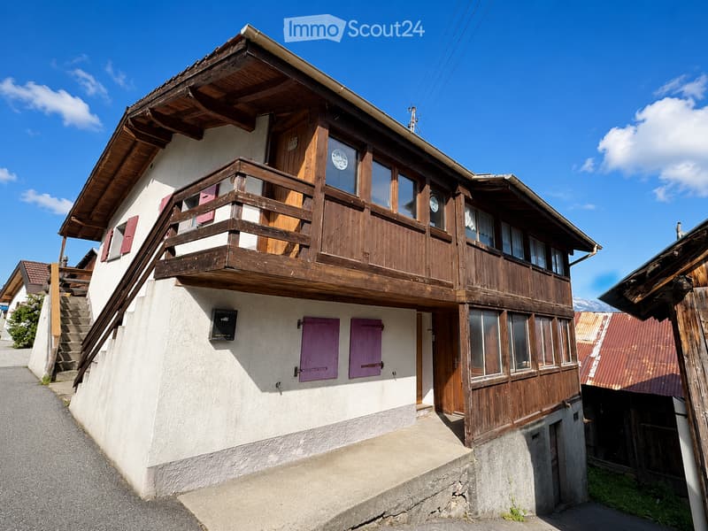 Two-story house with a wooden balcony, white walls, and purple shutters. The roof is wooden, and the house is situated on a raised ground floor with a staircase leading up to it.