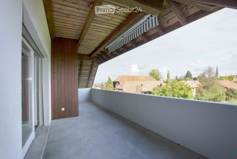 Balcony with wooden ceiling, grey concrete floor, grey railing, and a view of a town with houses and trees.