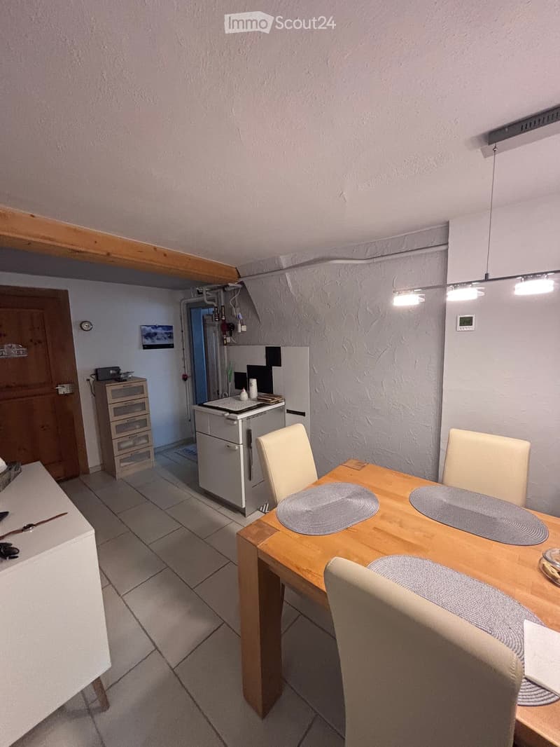 A small, well-lit kitchen and dining area with a wooden dining table, four chairs, a refrigerator, a small counter with a screen, and a drawer unit.