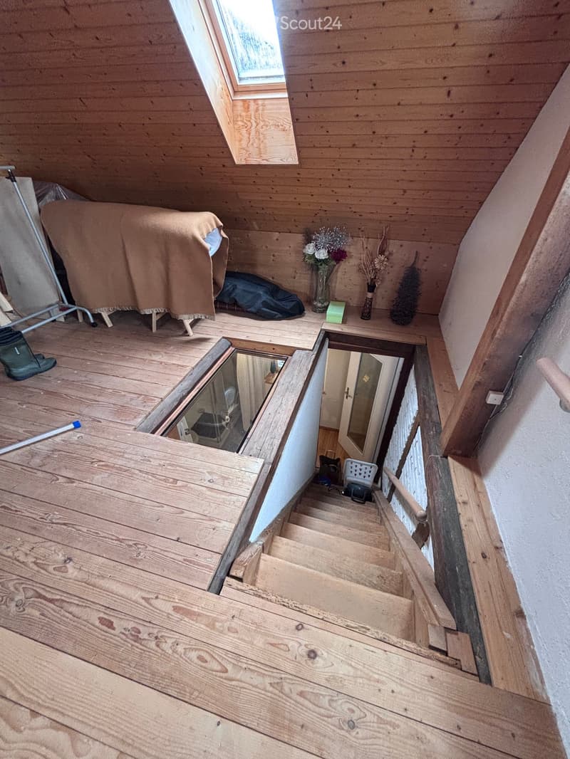 Wooden staircase leading up to an attic space with wooden walls and a skylight window. The attic contains a bed covered with a brown blanket, a small table with vases, and a mirror.