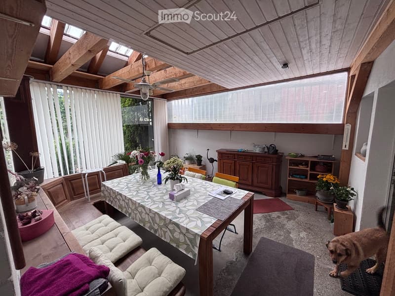 Brightly lit room with wooden beams, large windows, a dining table with chairs, a sofa, cabinets, and potted plants.