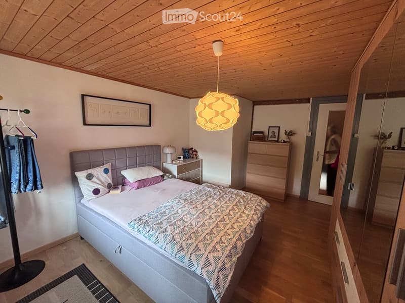 Cozy bedroom with a grey upholstered headboard, patterned bedding, a nightstand with a lamp, and a hanging coat rack. Wooden flooring and a modern pendant light are visible.