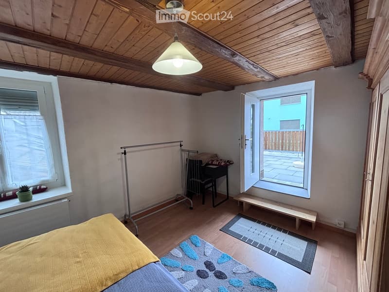 Cozy bedroom with wooden ceiling beams, a bed with a yellow blanket, a small table with a chair, a hanging lamp, and an open window leading to a patio.