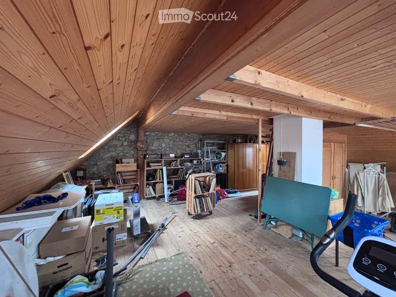 An attic space with wooden beams and exposed wooden floor, featuring a table, shelves, an exercise bike, and various storage items.