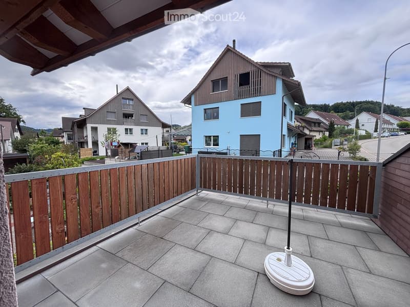Balcony with wooden railing, tiled floor, and a white umbrella base, overlooking a residential area with houses and greenery.