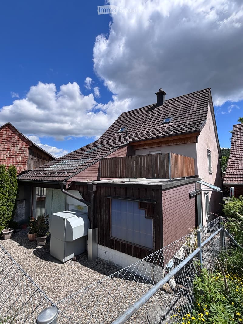 Two-story house with a tiled roof, wooden balcony, and surrounding greenery.