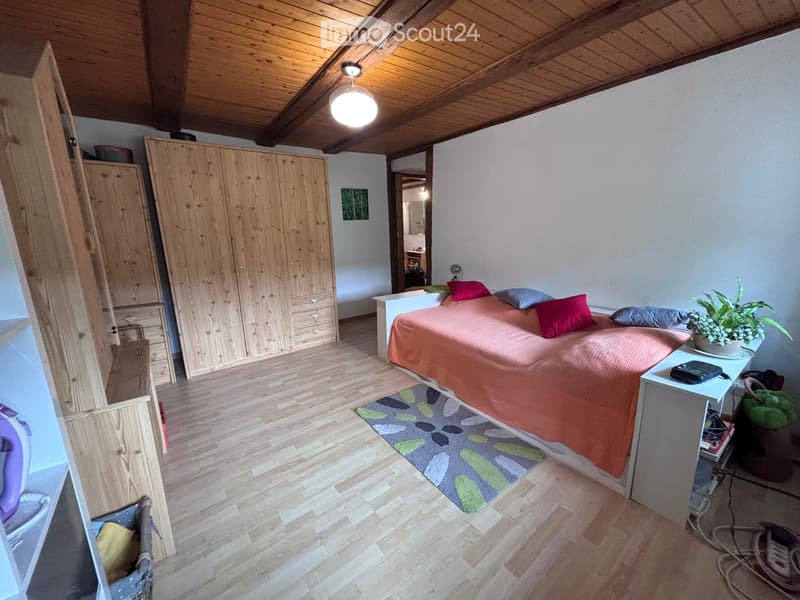 Cozy bedroom with wooden floors, slanted wooden ceiling, bed with orange bedding, nightstand with lamp, potted plant, and large wooden wardrobe.
