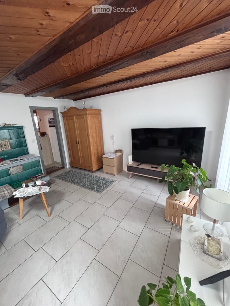 Living room with tiled floor, exposed wooden ceiling beams, a sofa, coffee table, TV, cabinet, and decorative plant.
