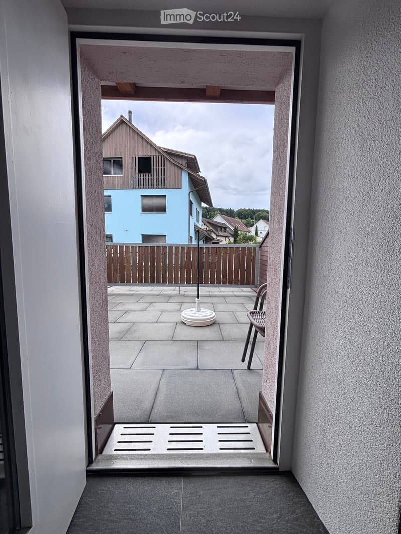 Balcony with tiled floor, white umbrella base, and a single chair, overlooking a residential area with houses and a wooden fence.