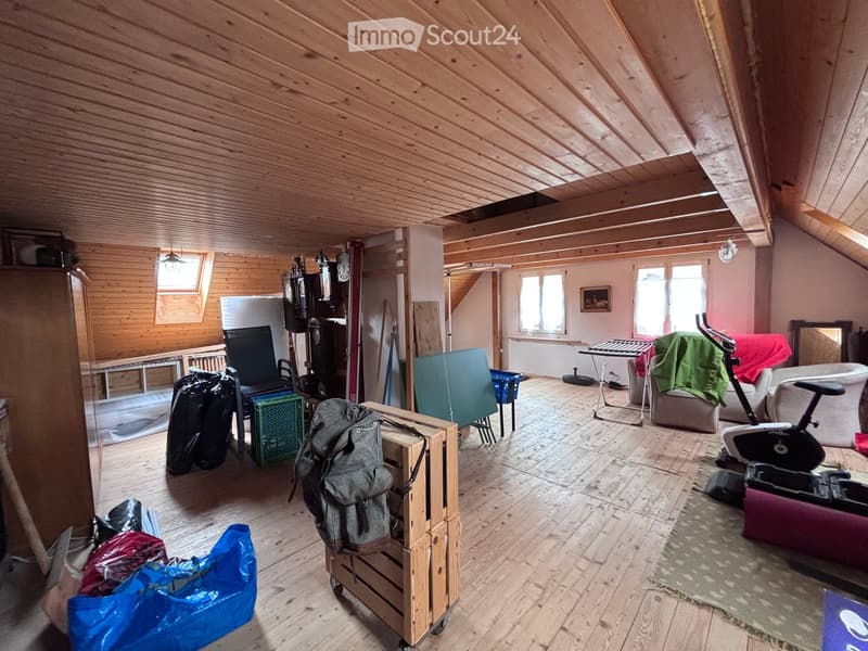 An attic room with wooden beams and flooring, featuring a sofa, chair, backpack, table, clock, mirror, and exercise bike.