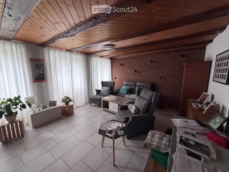 Living room with exposed wooden ceiling beams, brick accent wall, tiled floor, and large windows with white curtains. Features a grey sofa, armchairs, coffee table, side tables, and decorative plants.