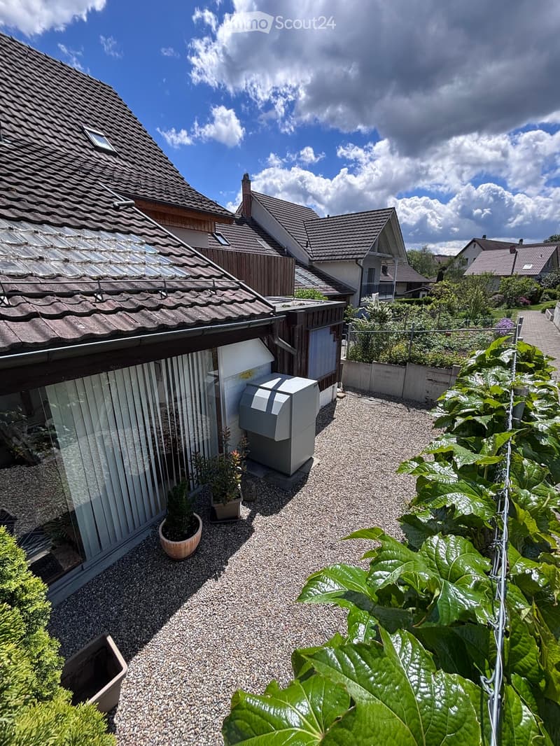 Garden area with gravel pathway, potted plants, and surrounding houses with tiled roofs.