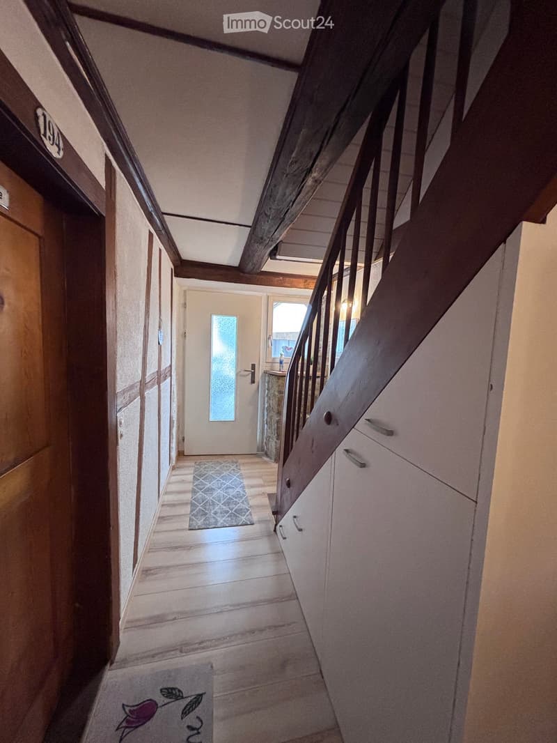A narrow hallway with wooden flooring, a small patterned rug, and a staircase leading upwards. The walls are light-colored, and there are doors on either side.